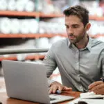 Warehouse Manager Taking Notes And Checking Orders With A Laptop