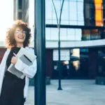 Lovely Curly Haired Entrepreneur With Glasses Smiling Outside While Posing With A Laptop And Tablet