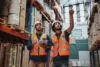 Young Caucasian Worker Showing Shelves To His African Manager In A Warehouse Holding Digital Tablet With Hardhat And Safety Vest