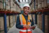 Happy Male Factory Manager Using Digital Tablet In Warehouse While Standing Against Goods Shelf Looking At Camera