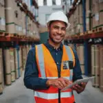 Happy Male Factory Manager Using Digital Tablet In Warehouse While Standing Against Goods Shelf Looking At Camera