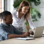 Smiling Businesswoman Mentor Helping New African American Employee With Software