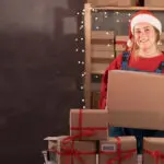 A Young Woman Wearing A Santa Claus Hat Works In A Warehouse Of Boxes With Gifts And Orders From An Online Store For Christmas. Small Business Owner Sells Happy New Year. Delivery Holiday.