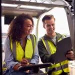 Male And Female Staff Operating Fork Lift Truck In Modern Warehouse