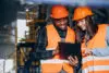 Three Factory Workers In Safety Hats Discussing Manufacture Plan