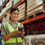 Phone, Engineer And Woman In Warehouse For Shipping, Distribution Or Stock Inspection On App. Mobile, Logistics And Happy Factory Worker Check Inventory For Export, Supply Chain And Quality Control