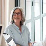 Happy Old Female Mentor Talking To Young Interns At Group Office Meeting.