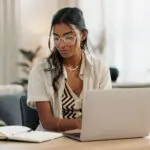 Woman With Laptop, Notes And Typing In Home Office Planning Freelance Research Project At Desk. Computer, Internet Search And Thinking, Girl With Glasses Writing Online Article For Remote Work Job.