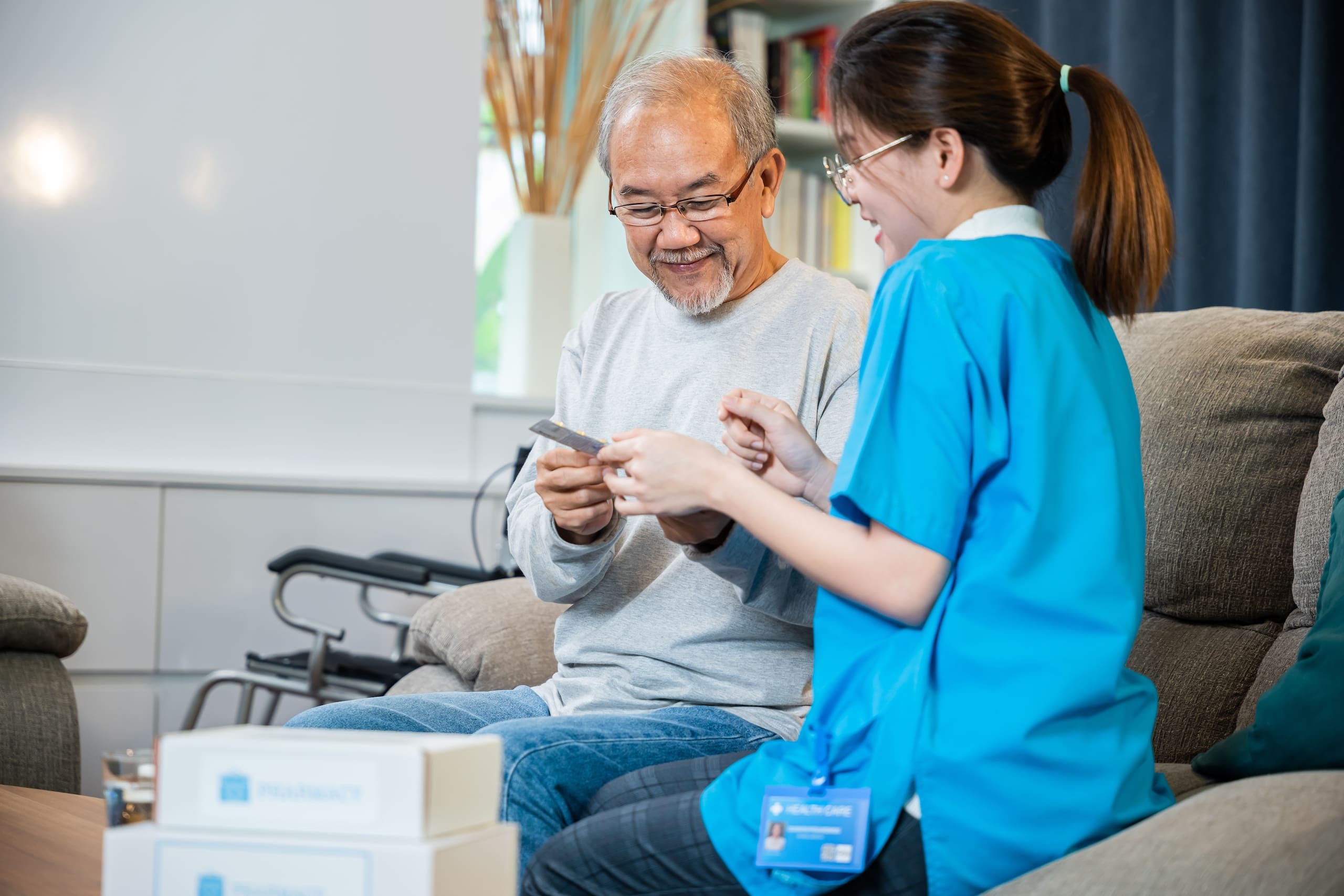 Asian Nurse With Physician Explaining Prescription Medicine To Attentive Senior Man At Home