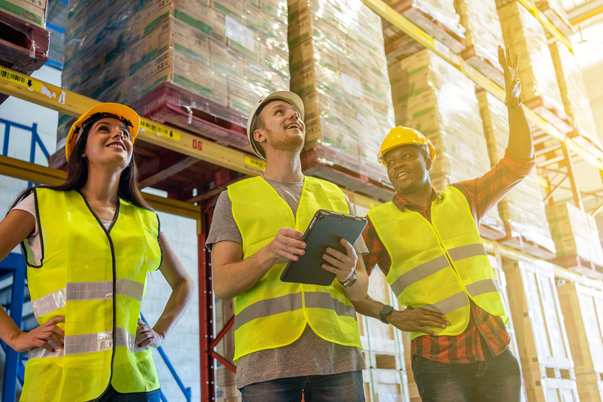 Warehouse Workers Team Meeting Using Tablet Computer With Training Work In Safety Helmet Standing In Storage Factory.warehouse Manager And Worker Working In Large Warehouse With Goods Happy