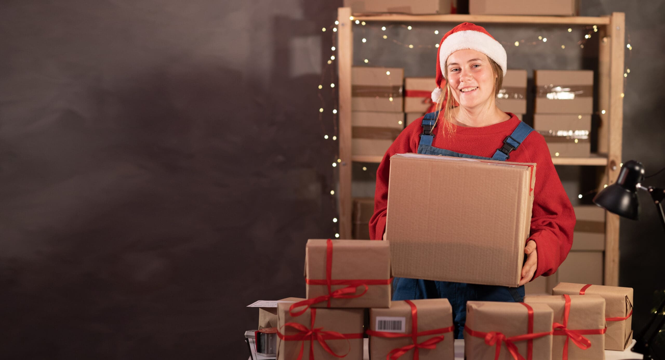 A Young Woman Wearing A Santa Claus Hat Works In A Warehouse Of Boxes With Gifts And Orders From An Online Store For Christmas. Small Business Owner Sells Happy New Year. Delivery Holiday.