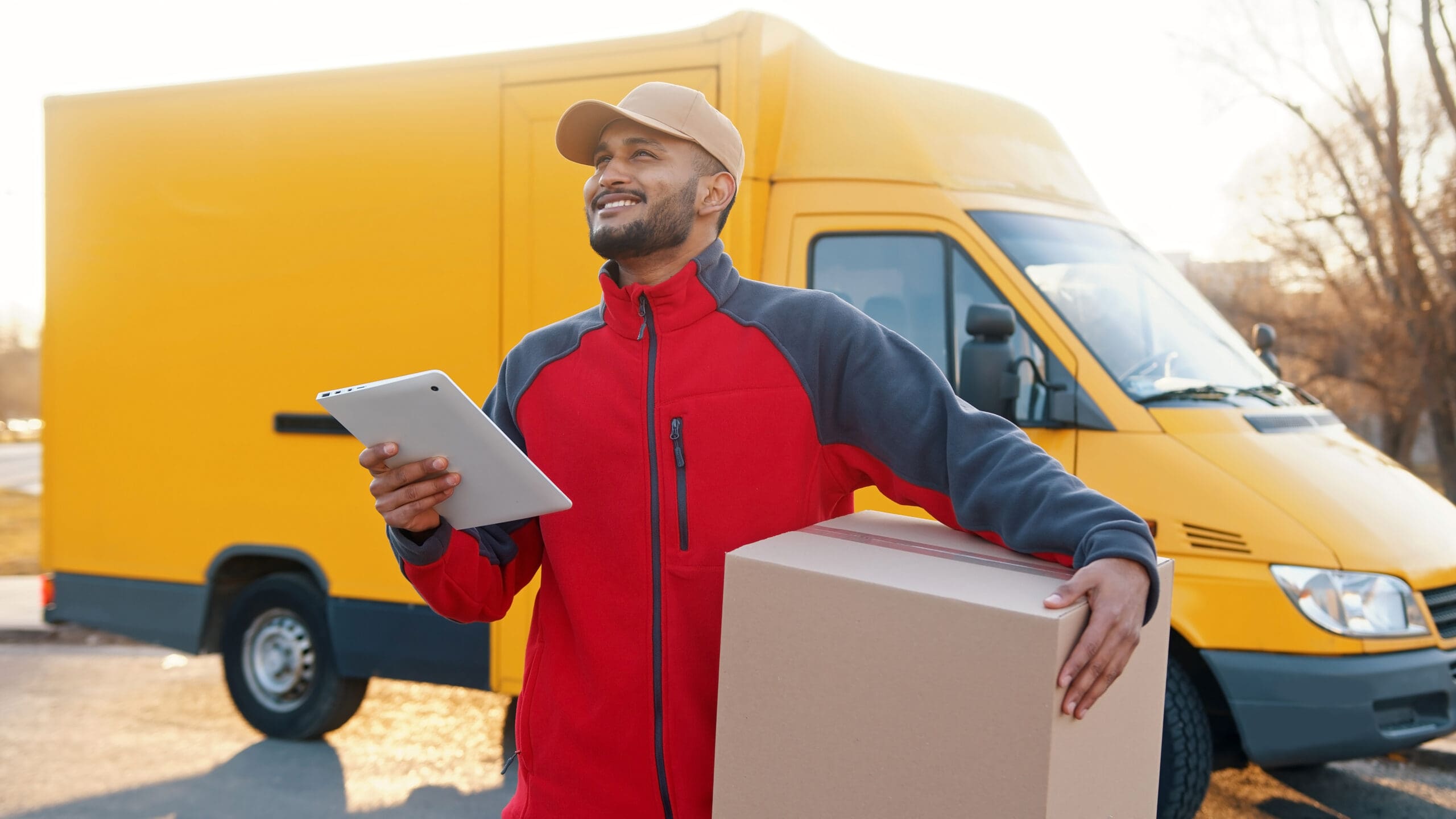 Delivery Boy With A Cardboard Box And An Ipad A Yellow Van Parked Behind Him